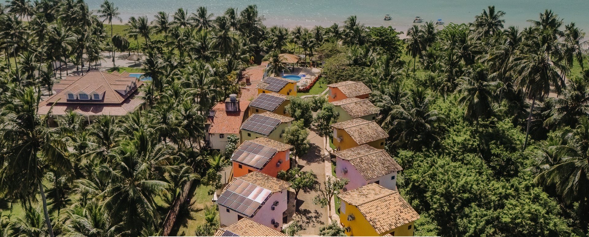 Vista aérea dos chalés coloridos da Pousada Residencial Maragogi entre coqueiros, com o mar de Ponta de Mangue ao fundo